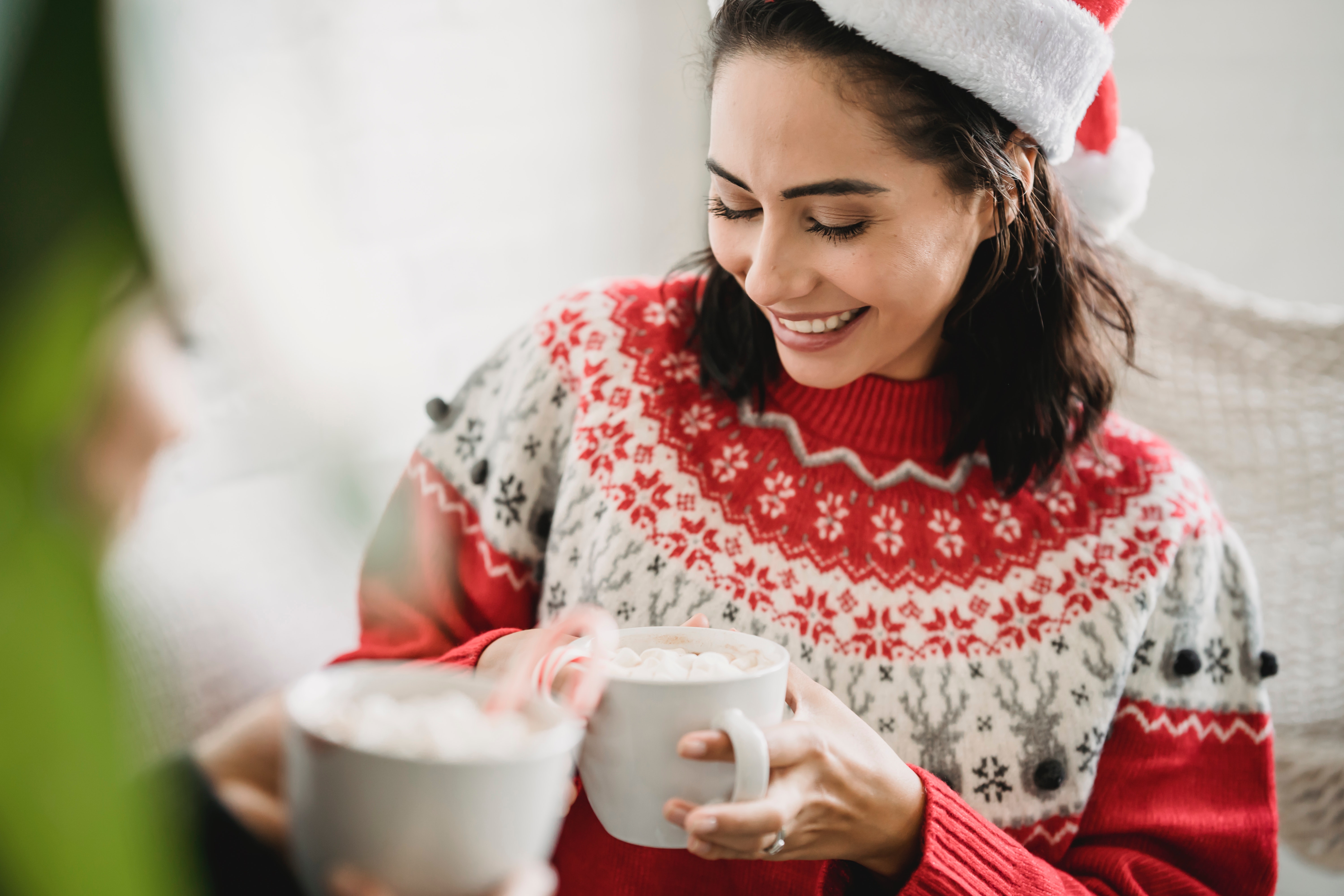 A woman in a red holiday sweater and a Santa hat drinking hot chocolate with a candy cane in it.